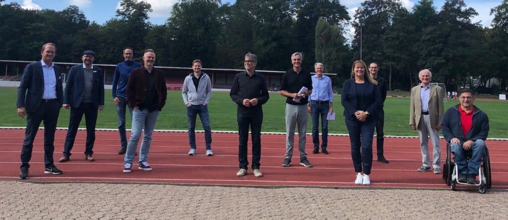 Gruppe von 13 Personen steht auf einer Laufbahn eines Sportplatzes, eine Person sitzt im Rollstuhl.