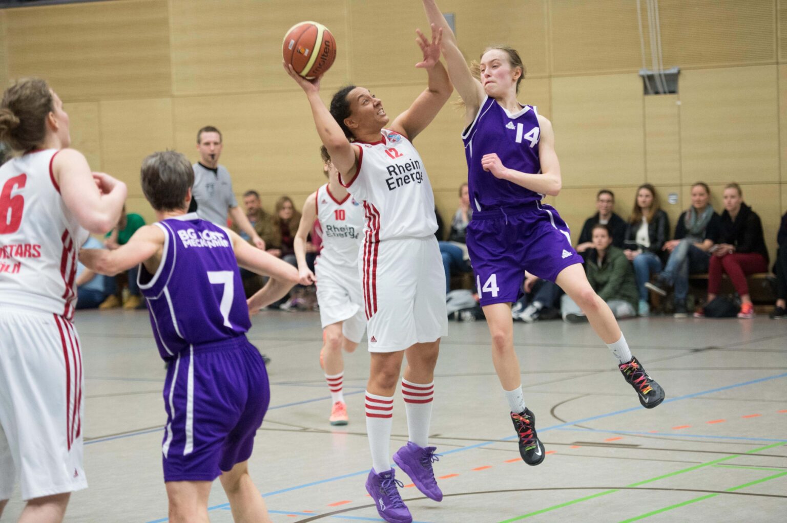 Basketballspiel der RheinStars Köln in der Halle: Spielerin im weißen Trikot wirft auf den Korb, während Gegnerinnen in Lila verteidigen.
