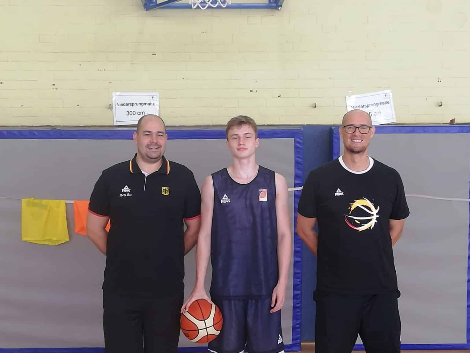 Drei Personen posieren in einer Sporthalle: zwei Trainer der RheinStars Köln und ein Basketballspieler mit Ball.