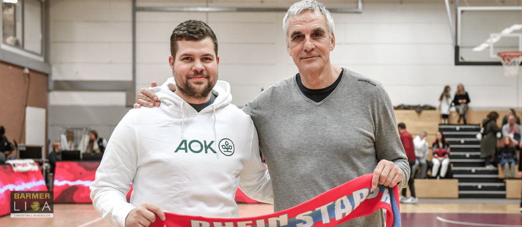 Zwei Männer halten ein rotes Banner in einer Sporthalle mit Zuschauern im Hintergrund.
