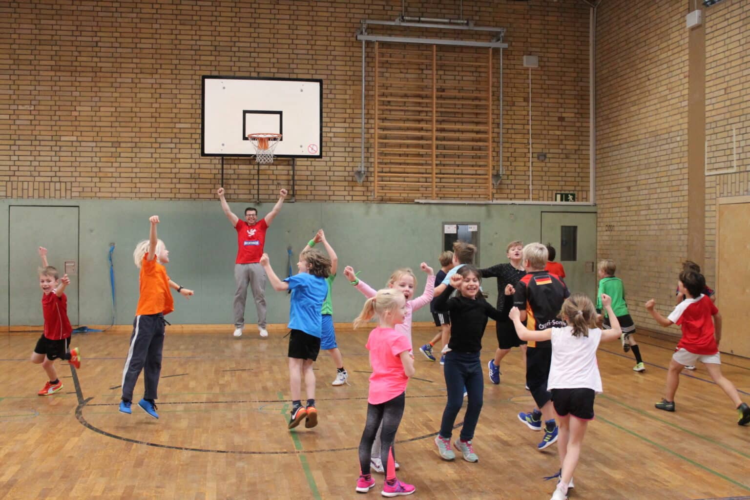 Gruppe von Kindern beim Basketball-Training in einer Sporthalle mit Basketballkorb an der Wand.