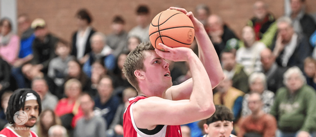 Basketballspieler der RheinStars Köln wirft Ball von außen, Zuschauer im Hintergrund.
