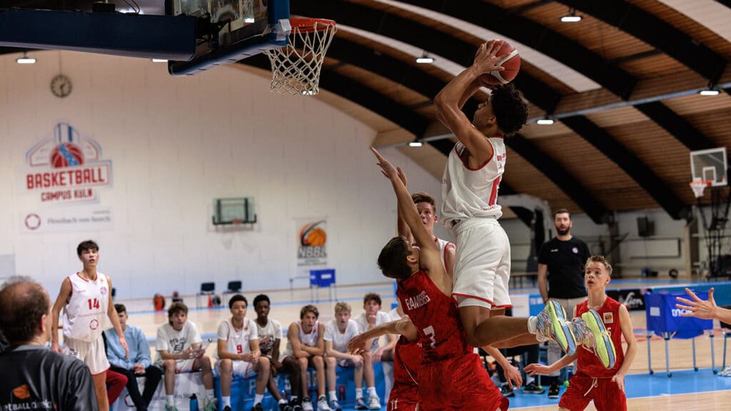 Basketballspiel der RheinStars Köln: Spieler in Weiß springt zum Wurf, Gegenspieler in Rot versucht zu blocken.