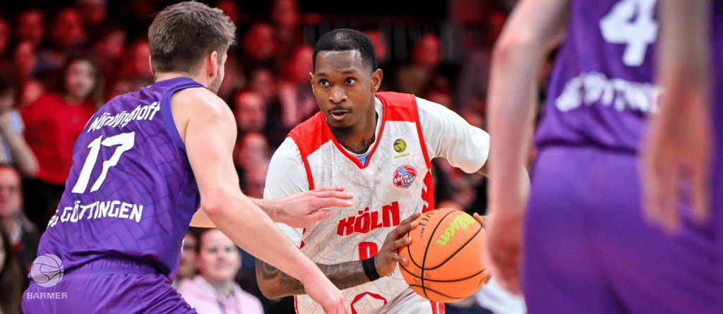 Basketballspieler der RheinStars Köln im roten Trikot dribbelt gegen Verteidiger im violetten Trikot.