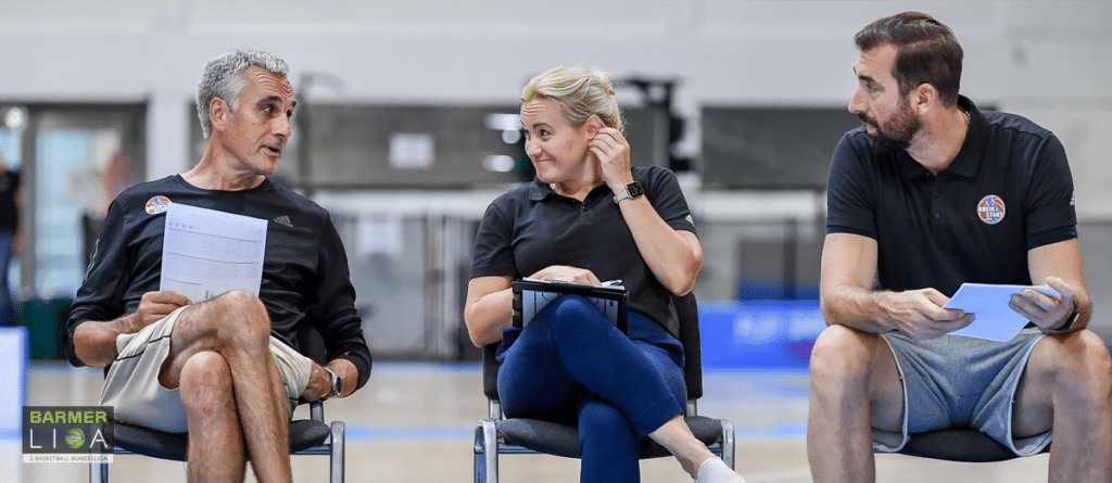 Drei Personen sitzen in einer Sporthalle der RheinStars Köln und diskutieren, einer hält ein Klemmbrett.