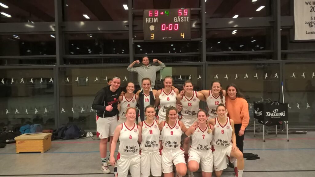 Frauenbasketballteam RheinStars Köln posiert gemeinsam in der Sporthalle vor dem Scoreboard.