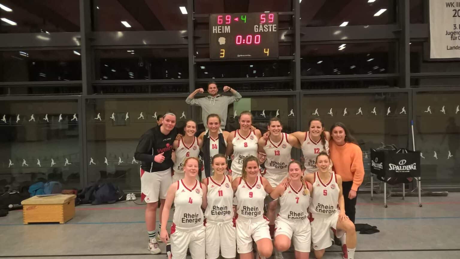 Frauenbasketballteam RheinStars Köln posiert gemeinsam in der Sporthalle vor dem Scoreboard.