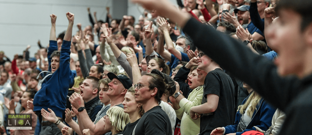 Enthusiastische Fans der RheinStars Köln jubeln mit erhobenen Armen in einer Sporthalle.
