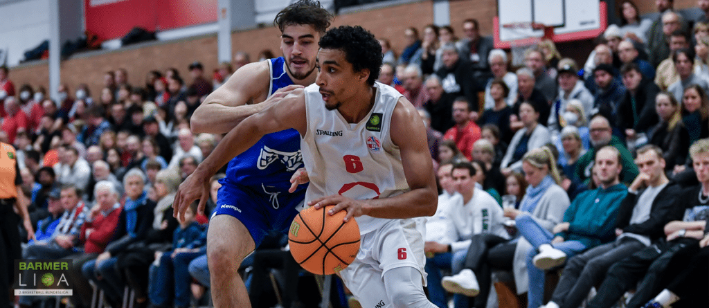 Basketballspieler der RheinStars Köln dribbelt Ball, verteidigt von Gegenspieler in blauem Trikot.