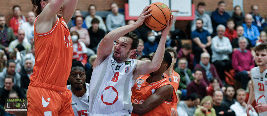 Basketballspiel der RheinStars Köln: Spieler in Weiß mit Ball umgeben von Gegnern in Orange.