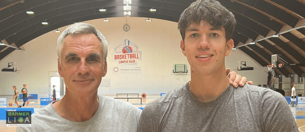 Zwei Männer in einer Basketballhalle mit RheinStars Köln Logo im Hintergrund.