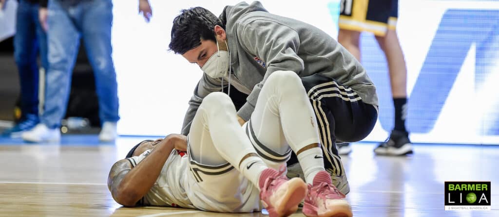 Basketballspieler der RheinStars Köln liegt verletzt auf dem Hallenboden, Betreuer kümmert sich um ihn.
