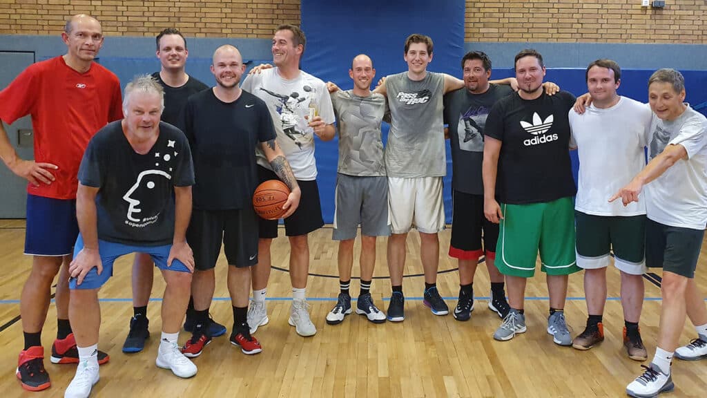 Elf Männer der RheinStars Köln posieren lächelnd in einer Sporthalle mit Basketball.