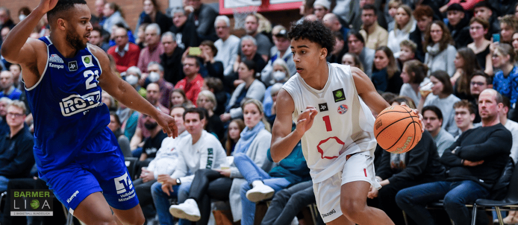 Basketballspieler der RheinStars Köln dribbelt gegen Gegenspieler vor Zuschauern.