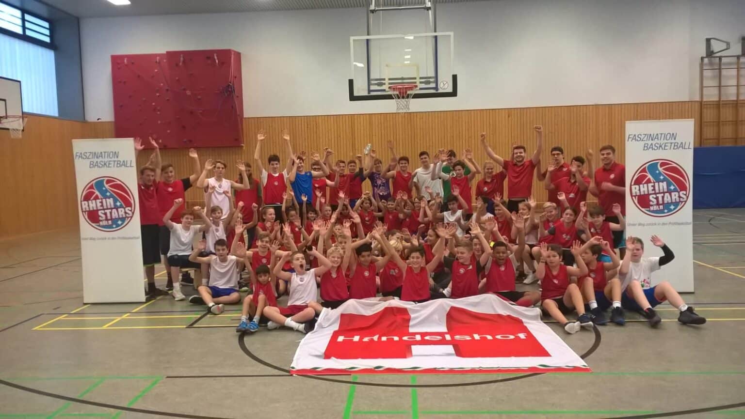 Große Gruppe junger Basketballer der RheinStars Köln posiert jubelnd in einer Sporthalle mit Flagge.