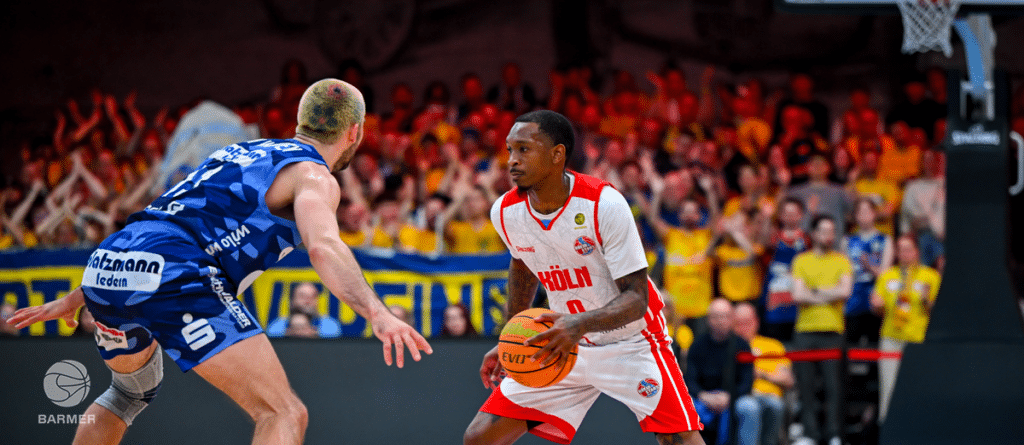 Basketballspiel der RheinStars Köln: Spieler in Rot-Weiß dribbelt gegen Verteidiger in Blau, Publikum im Hintergrund.