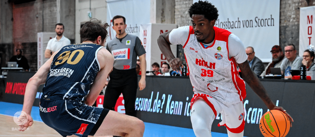 Basketballspiel der RheinStars Köln gegen Eisenbahn Strasbourg, Spieler in Rot-Weiß mit Ball gegen Verteidiger in Dunkelblau.