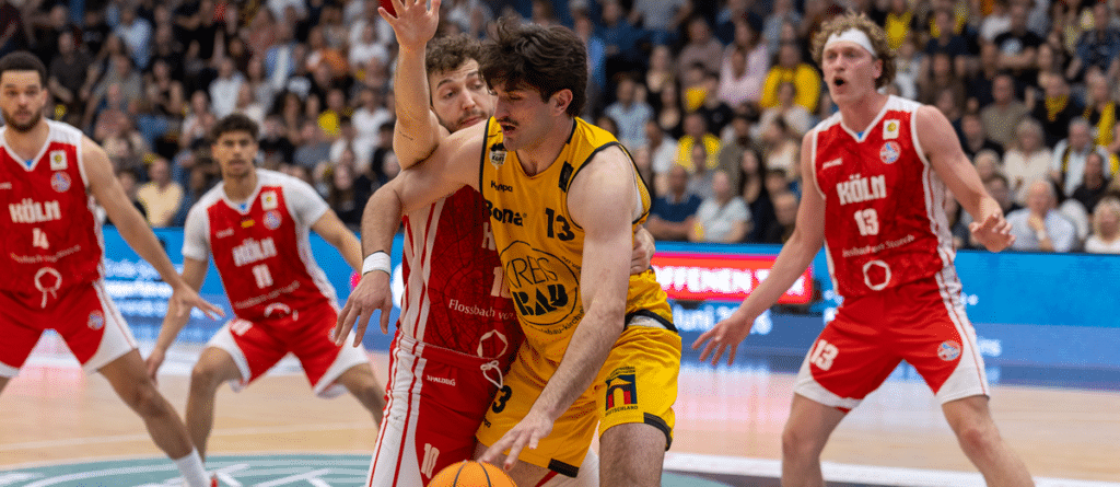 Basketballspiel der RheinStars Köln gegen Team in Gelb, Spieler mit Ball umgeben von Gegnern in Rot.