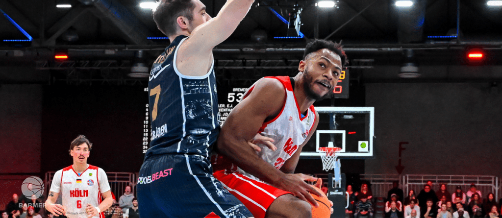 Basketballspiel der RheinStars Köln: Spieler in rotem Trikot dribbelt gegen Gegner in blauem Trikot in vollbesetzter Halle.