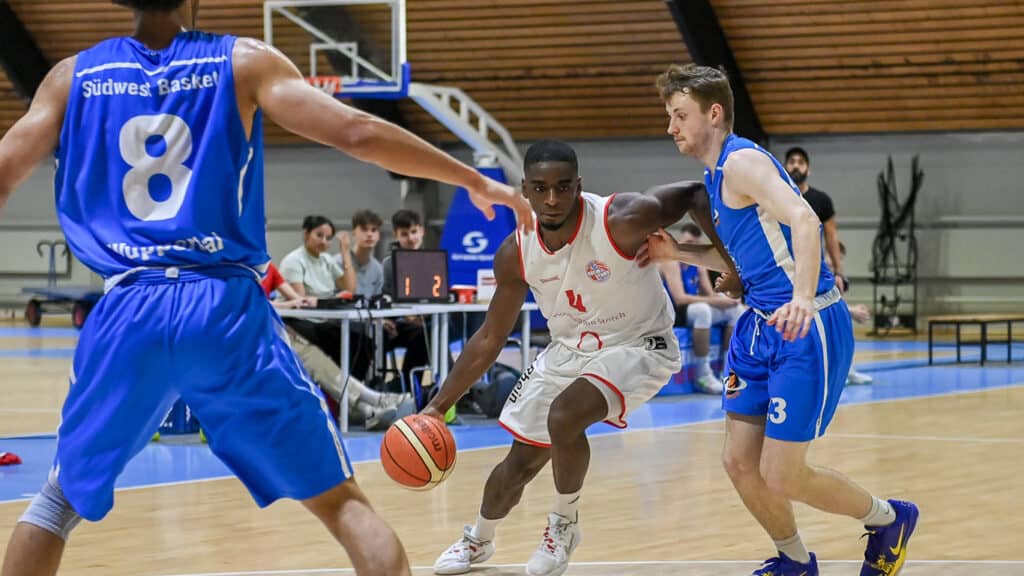 Basketballspiel: RheinStars-Spieler in Weiß dribbelt gegen Südwest-Basketballer in Blau.