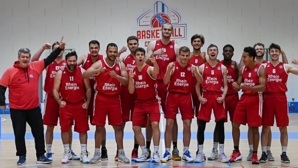 Basketballteam RheinStars Köln posiert jubelnd in roter Trainingskleidung in einer Sporthalle.