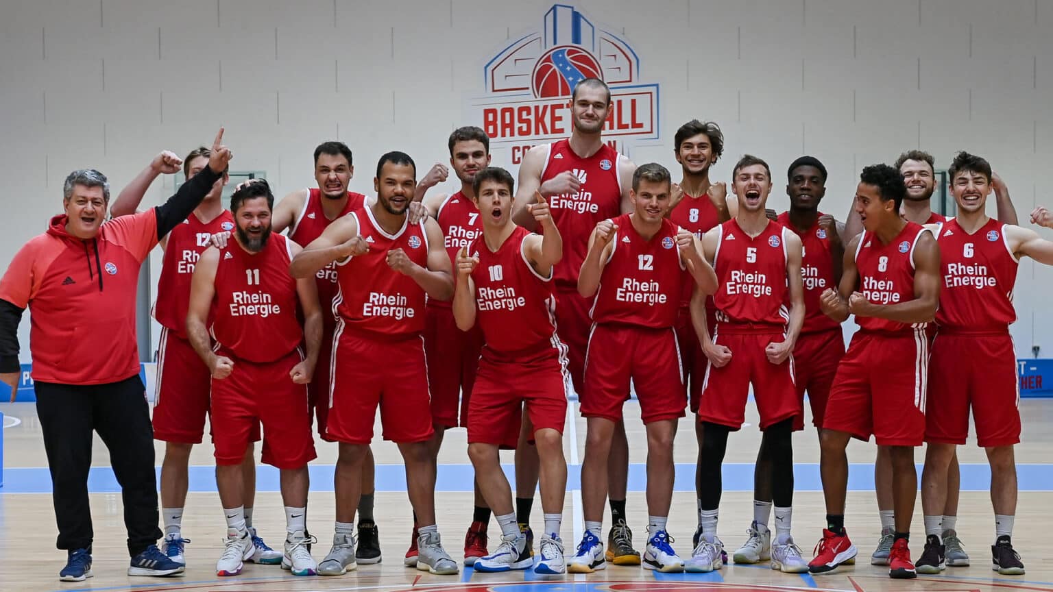 Basketballteam RheinStars Köln posiert jubelnd in roter Trainingskleidung in einer Sporthalle.