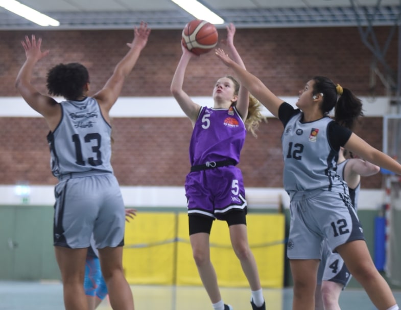 Basketballspiel der RheinStars Köln: Spielerin in Lila wirft Ball, zwei Gegnerinnen in Grau verteidigen.
