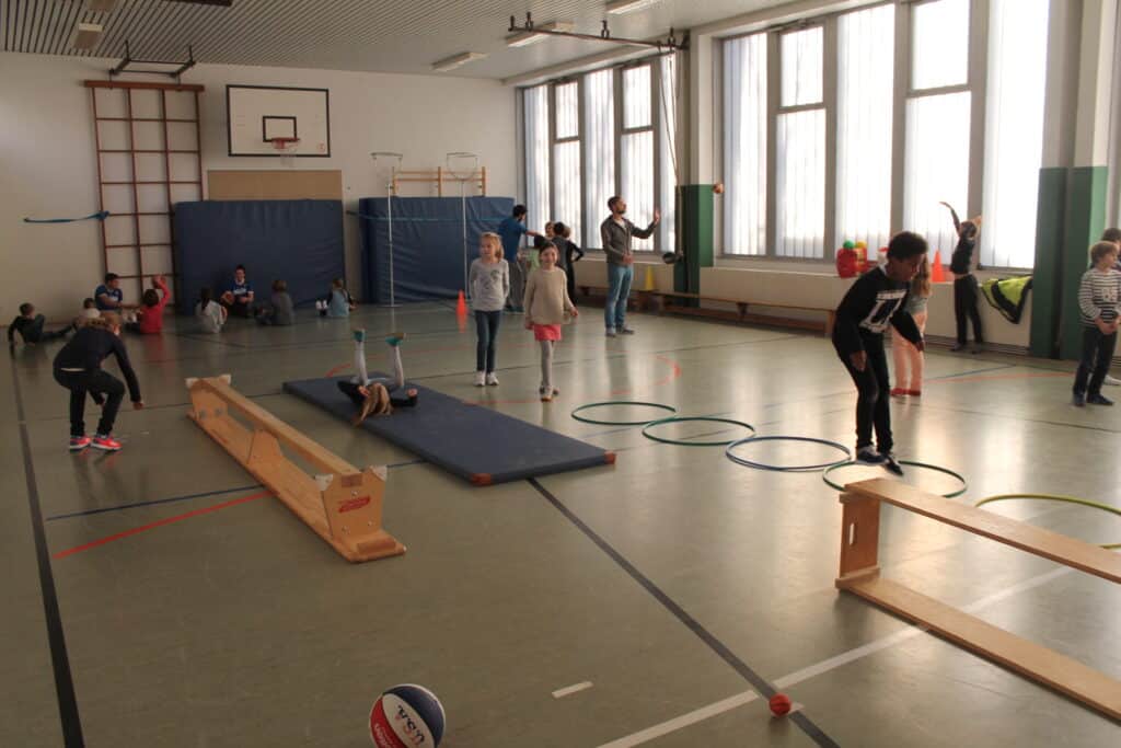 Sporthalle mit Kindern bei Bewegungsübungen der RheinStars Köln; Geräte wie Balken, Matten und Ringe sind aufgebaut.