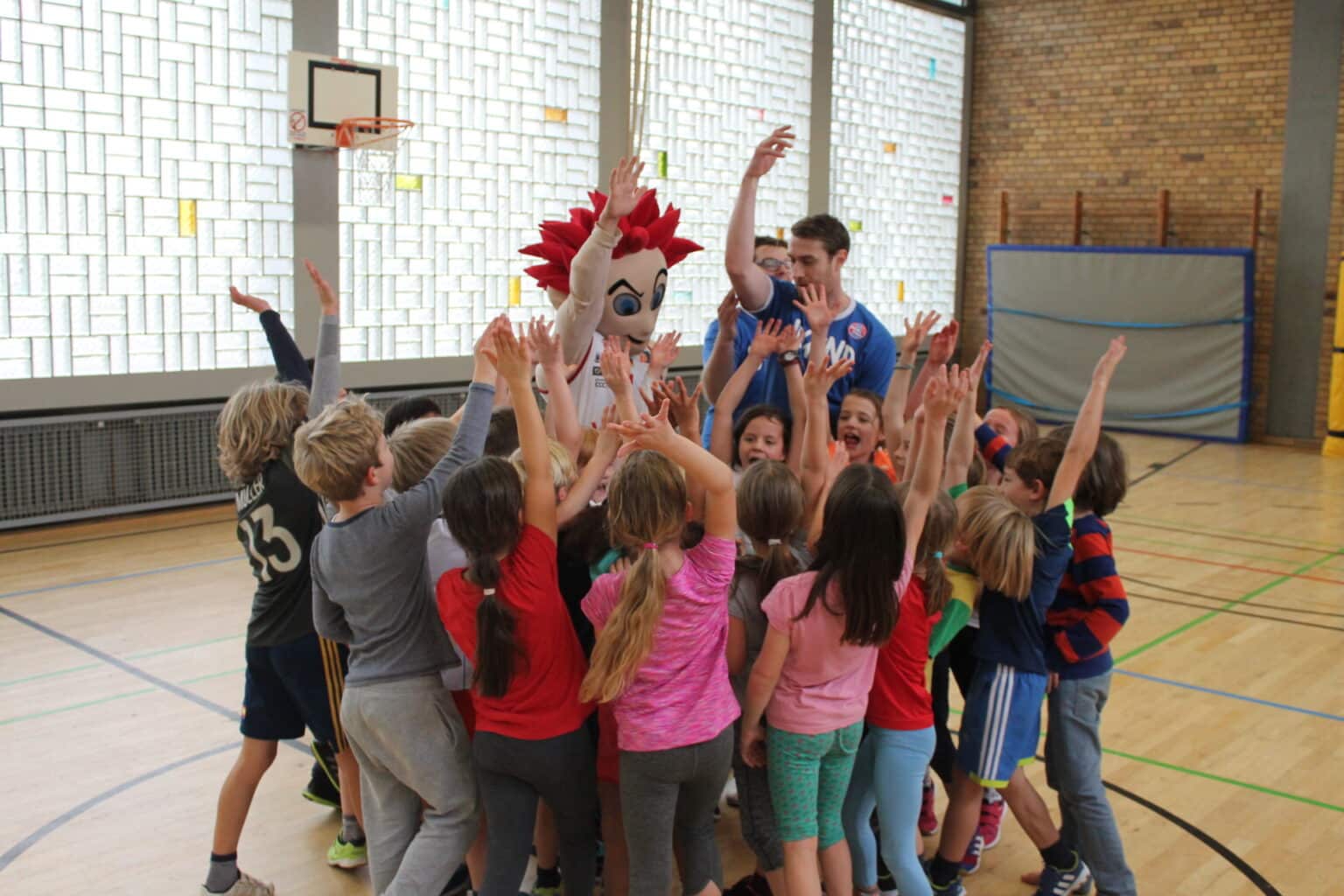 Kinder jubeln in einer Sporthalle um einen Maskottchen-Charakter mit roten Haaren herum.