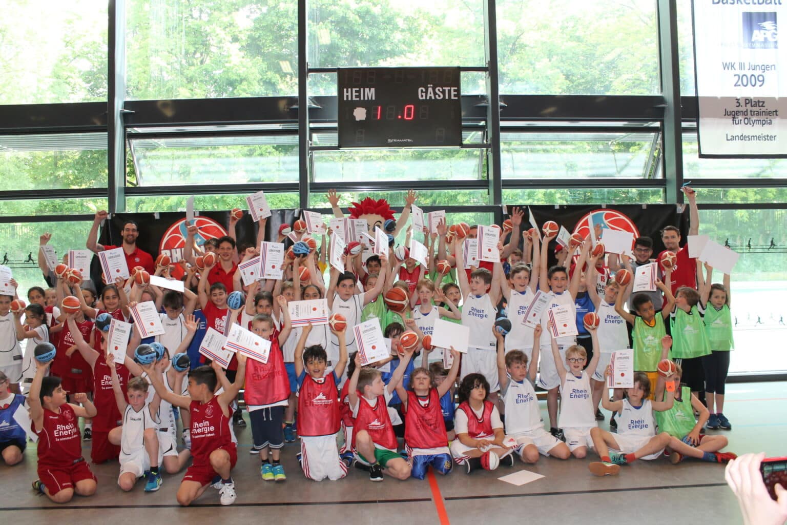 Große Gruppe junger Basketballspieler der RheinStars Köln posiert mit Urkunden und Basketbällen in einer Sporthalle.