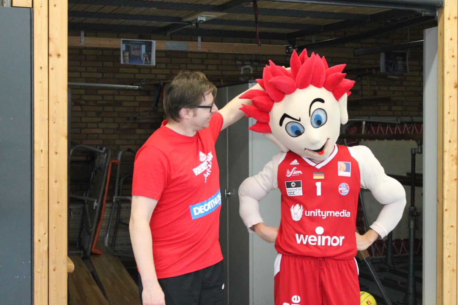 Ein Mann in rotem Decathlon-Shirt posiert mit dem Maskottchen der RheinStars Köln in einer Sporthalle.