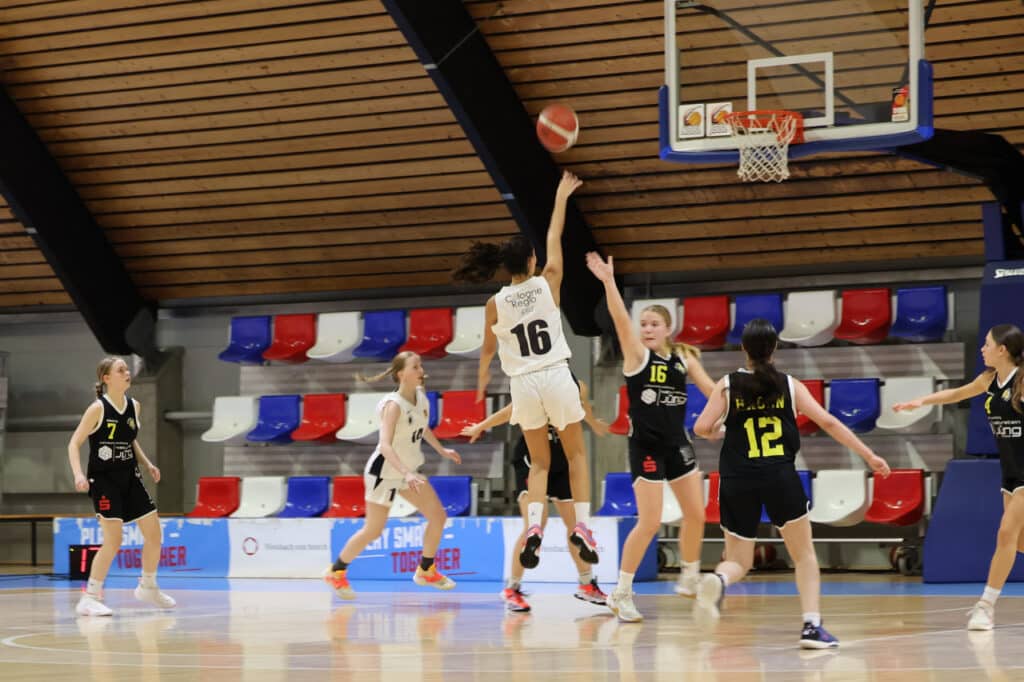 Basketballspiel der RheinStars Köln: Spielerin Nummer 16 wirft Ball auf Korb, Gegnerinnen verteidigen in schwarzen Trikots.