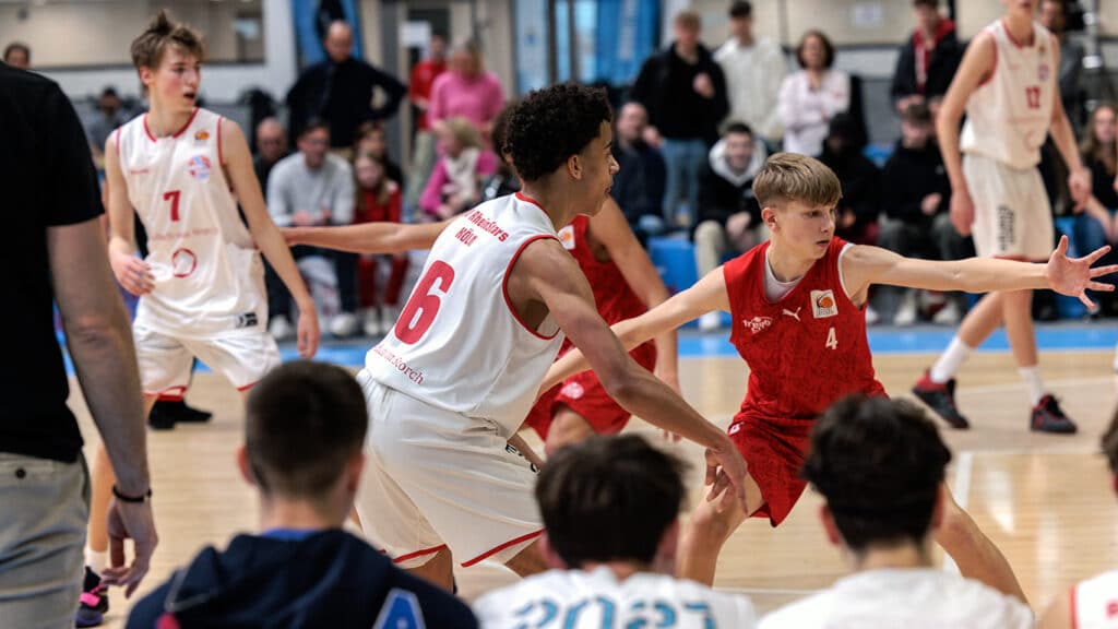 Basketballspiel der RheinStars Köln: Spieler Nummer 6 in weißem Trikot verteidigt gegen Gegner in rotem Trikot vor Zuschauern in der Halle.
