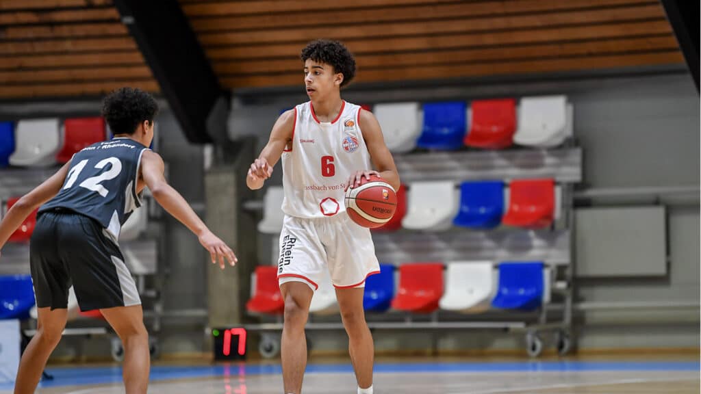 Basketballspiel der RheinStars Köln: Spieler Nummer 6 im weißen Trikot dribbelt gegen Gegner Nummer 12 in dunklem Trikot.