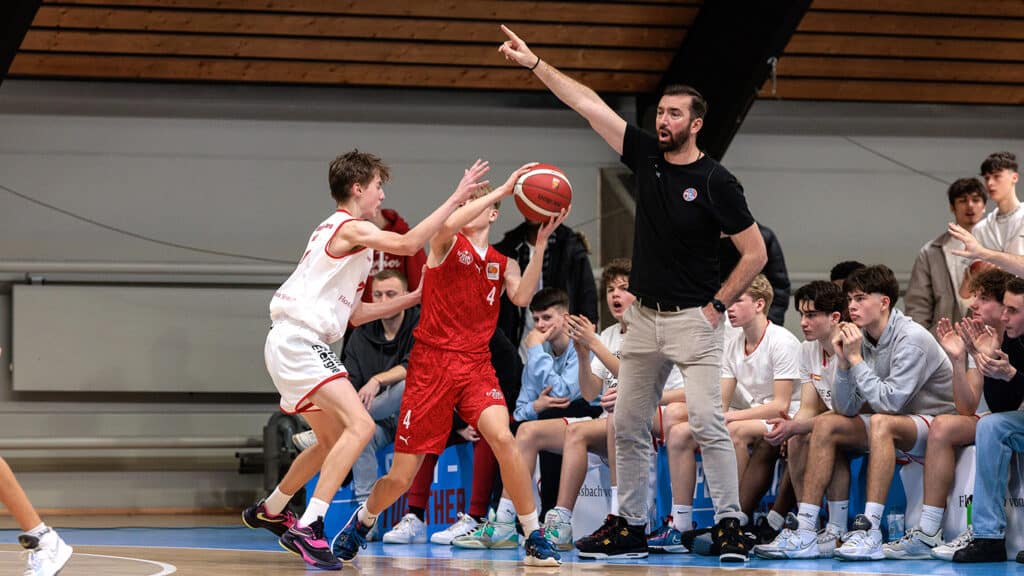 Basketballspiel der RheinStars Köln: Spieler in rotem Trikot wird von Gegenspieler in Weiß verteidigt, Trainer und Zuschauer an der Seitenlinie.