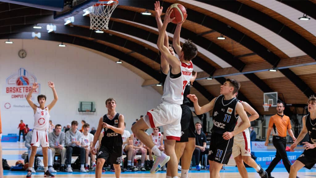 Basketballspiel der RheinStars Köln: Spieler in Weiß-Rot springt zum Wurf, Gegner in Schwarz verteidigt.