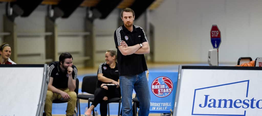 Mann in schwarzem Adidas-Shirt steht mit verschränkten Armen in einer Sporthalle der RheinStars Köln, drei weitere Personen sitzen daneben.