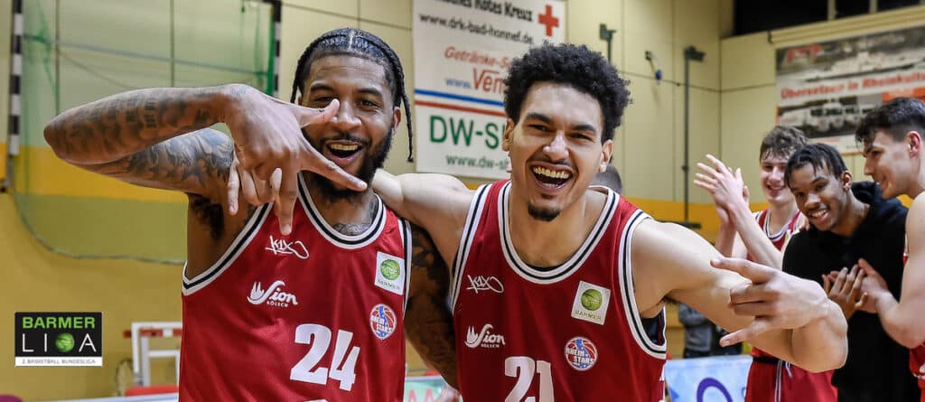 Zwei Basketballspieler der RheinStars Köln in roten Trikots feiern lachend in der Halle.