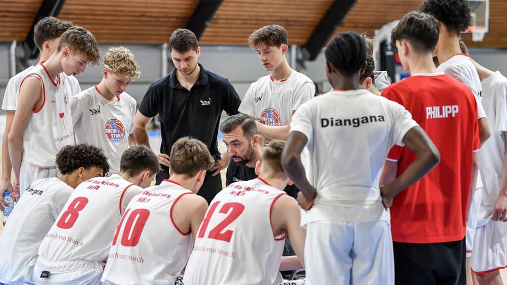 Trainer bespricht mit RheinStars-Köln-Basketballteam taktische Anweisungen in der Sporthalle.