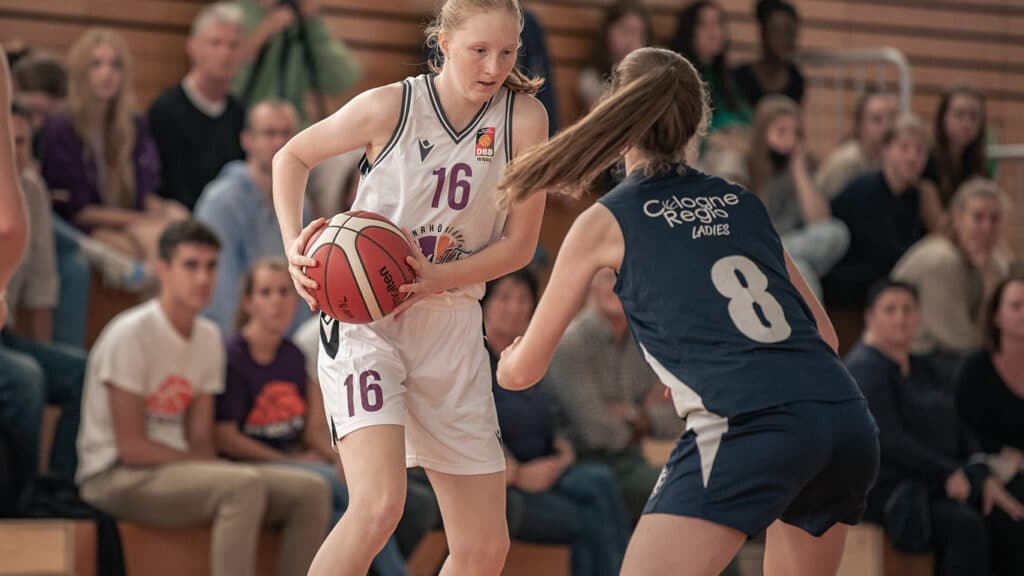 Basketballspielerin der RheinStars Köln Nummer 16 mit Ball gegen Abwehrspielerin Nummer 8 in einer Sporthalle.