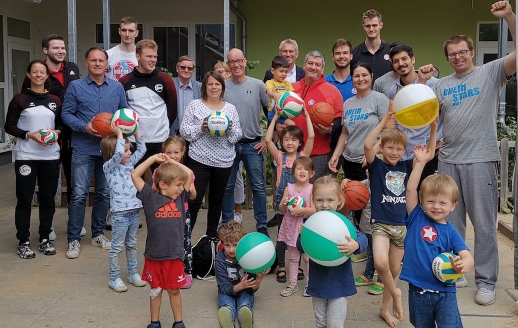 Große Gruppe von Erwachsenen und Kindern der RheinStars Köln mit Volleybällen beim Gruppenfoto vor grüner Wand.