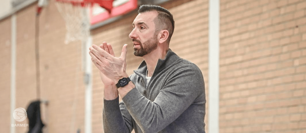 Mann in grauem Sportoberteil gestikuliert sprechend in einer Sporthalle der RheinStars Köln.