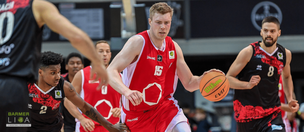 Basketballspiel der RheinStars Köln: Spieler Nummer 15 im roten Trikot dribbelt gegen Gegner in schwarzem Trikot.