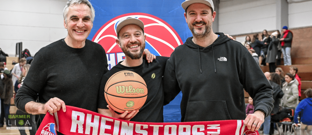 Drei Männer halten ein RheinStars-Köln-Banner und einen Basketball in einer Sporthalle.