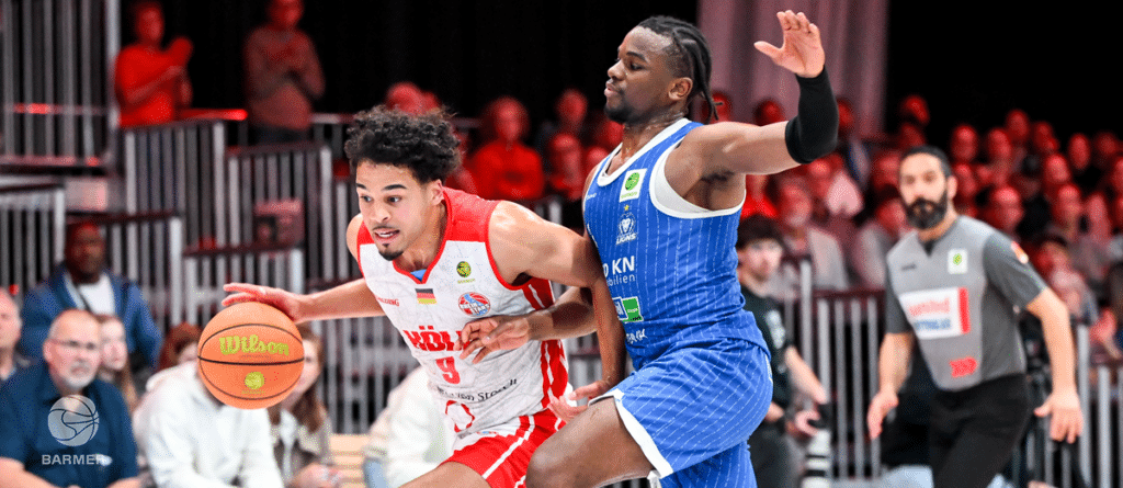 Basketball-Spieler der RheinStars Köln in rotem Trikot dribbelt gegen Verteidiger in blauem Trikot.