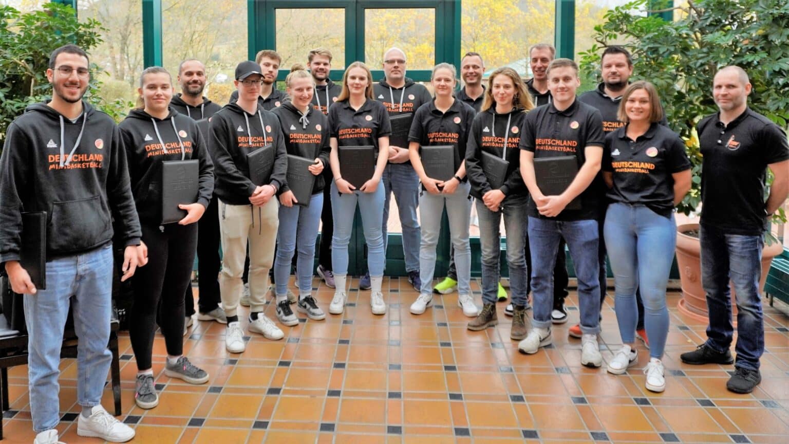 Mannschaftsfoto der RheinStars Köln: Etwa 18 Personen in schwarzen Team-Shirts mit "Deutschland" und "Minibasketball"-Aufdruck stehen auf einer Terrakotta-Fliesenfläche vor grünen Fenstern.