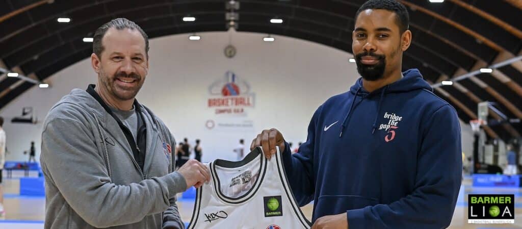 Zwei Männer halten ein signiertes Basketball-Trikot der RheinStars Köln in einer Sporthalle.