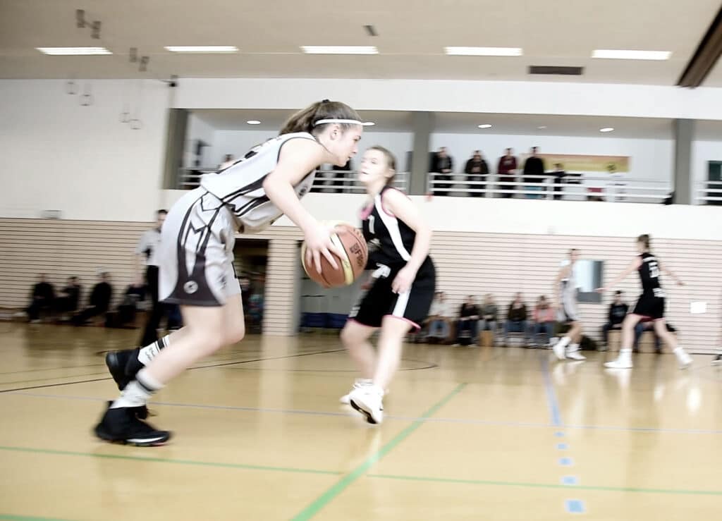 Basketballspiel der RheinStars Köln: Spielerin in weißem Trikot dribbelt gegen Verteidigerin in schwarzem Trikot in der Sporthalle.