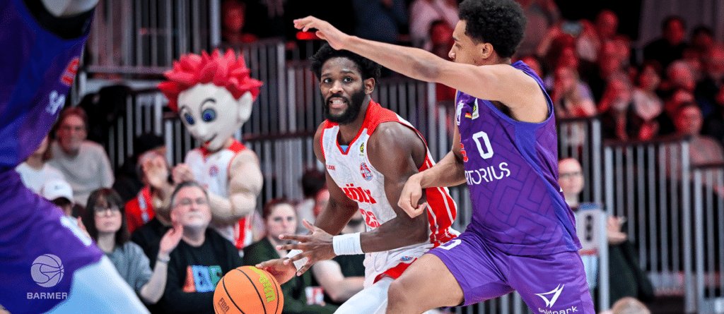 Basketballspiel: RheinStars-Spieler in Rot dribbelt gegen Gegner in Lila.