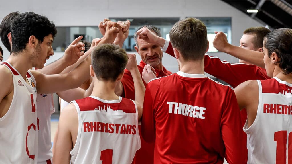 Basketballteam RheinStars Köln bei Mannschaftszeremonie mit Motivationsgeste im Kreis.